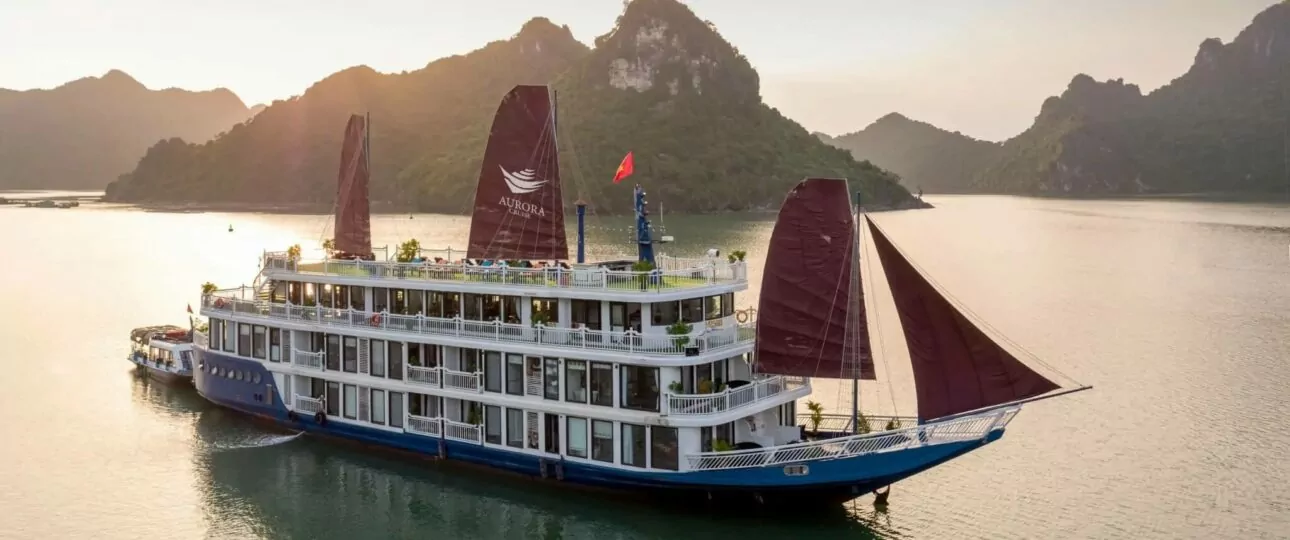 Aurora Cruise sailing Lan Ha Bay at sunset with three maroon sails raised against limestone karst mountains, Vietnam
