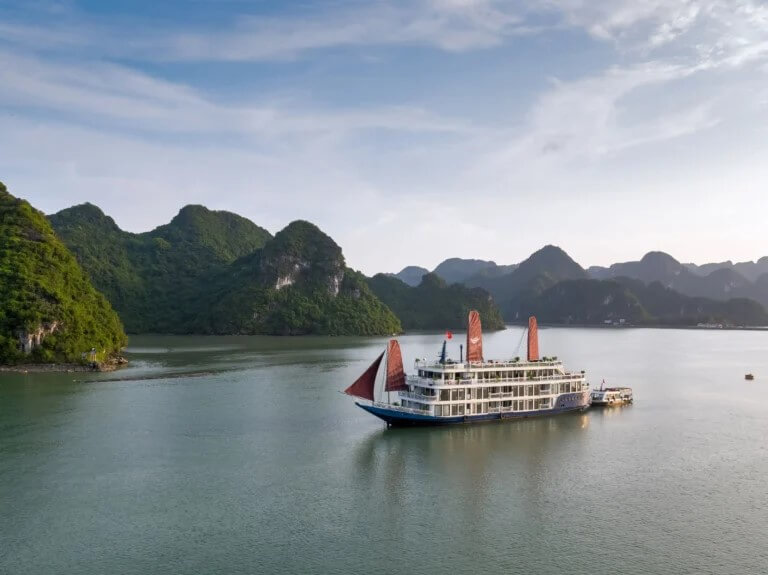Aurora Cruise anchored in Lan Ha Bay on a clear day, red sail raised, surrounded by limestone islands