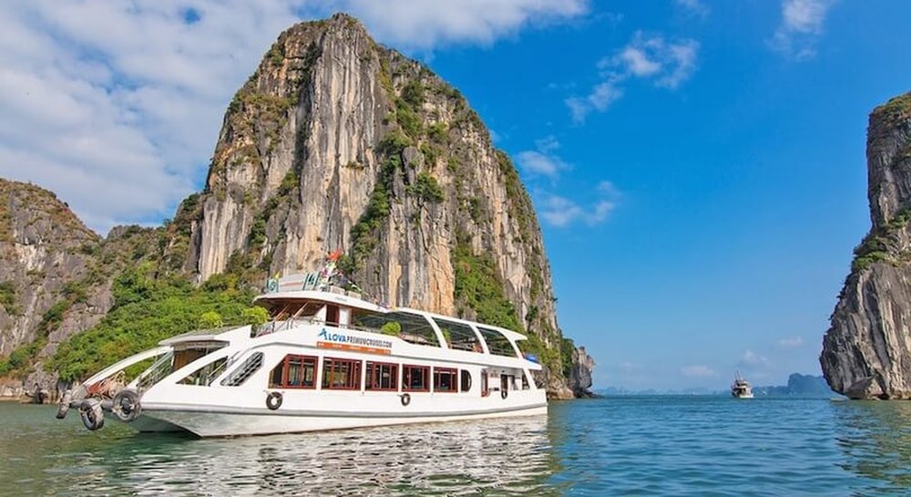 Alova Premium Day Cruise dwarfed by a towering limestone karst in Halong Bay